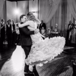 A groom lifts and spins his bride on the dance floor as guests watch at a wedding reception in a decorated venue.
