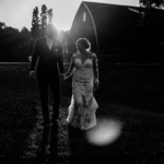 A couple in formal attire walks hand-in-hand outdoors at sunset, with a barn and trees in the background. The scene is in black and white.