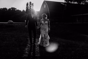 A couple in formal attire walks hand-in-hand outdoors at sunset, with a barn and trees in the background. The scene is in black and white.