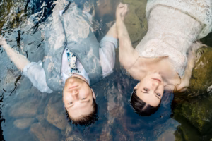 A man in a vest and tie and a woman in a lace dress lie side by side in shallow water, holding hands and looking up.