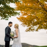 A bride and groom stand facing each other outdoors, holding a bouquet, with autumn trees and cloudy skies in the background.