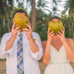 A man and a woman in formal attire hold coconuts in front of their faces while standing outdoors among palm trees.