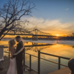 A bride and groom embrace near a railing at sunset by a river, with a bridge and bare trees in the background.