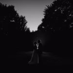A silhouetted couple stands close together outdoors at dusk, surrounded by dark trees and illuminated by fading light.