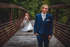 A bride in a white dress stands on a wooden bridge, looking back, while a man in a blue suit stands in the foreground out of focus. Lush trees surround the bridge.