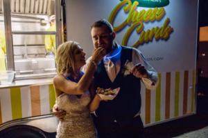 A bride feeds a groom cheese curds in front of a food truck at night; both are dressed in wedding attire and smiling.