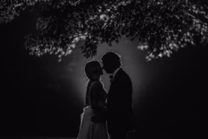 A couple stands close together under tree branches at night, facing each other in silhouette with soft backlighting.