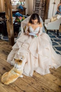 A bride in a white wedding dress sits indoors reading a note, while a small dog sits on the wooden floor in front of her.