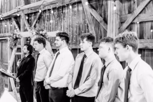 A group of six men dressed in formal shirts and ties stand in a row inside a rustic wooden venue with string lights and floral decorations.