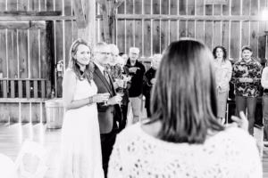 A group of people, including a woman in a white dress, stand indoors holding drinks and listening to a person speaking in the foreground.