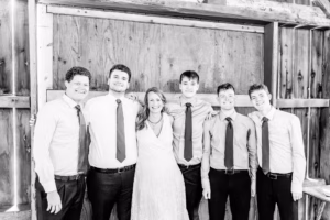 Five young men in dress shirts and ties stand with a woman in a white dress, smiling, against a wooden backdrop.