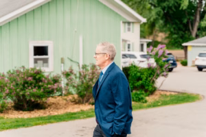 A man in a blue suit walks outside near a green house, with parked cars and greenery in the background.