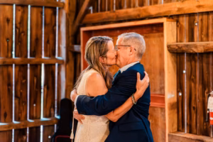 A woman in a white dress and an older man in a suit embrace and kiss inside a rustic wooden building.
