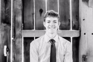 A young man wearing a dress shirt and tie sits in front of a wooden fence, smiling at the camera in a black and white photo.