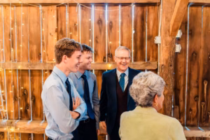 Three men in dress shirts and ties smile and converse with an older woman in a light sweater, standing in front of wooden walls with string lights.