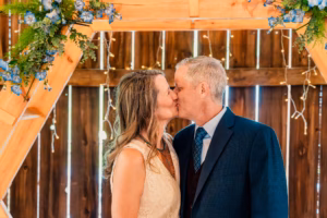 A couple dressed in formal attire kisses under a wooden arch decorated with blue flowers and greenery, with string lights in the background.