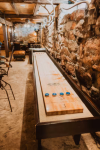 A shuffleboard table with blue pucks stands in a rustic room with stone walls, wooden ceiling, and vintage furniture.