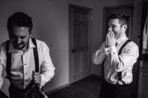 Two men in dress shirts and suspenders stand indoors; one is smiling with hands over his mouth, while the other looks down and holds a bag.