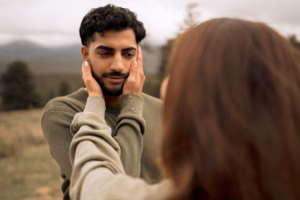 A person gently holds another person's face with both hands outdoors, with blurred trees and hills in the background.