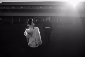 A bride in a white gown and a man in a suit walk across a field toward a large barn as the sun sets behind the building.