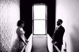 A bride and groom face each other across a stair landing, with a bright window between them in a black-and-white setting.