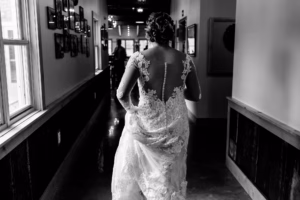 A person in a lace wedding dress walks down a hallway, photographed from behind in black and white.