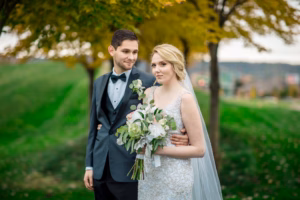 A groom in a tuxedo stands with his arm around a bride holding a bouquet, both posing outdoors with green grass and trees in the background.