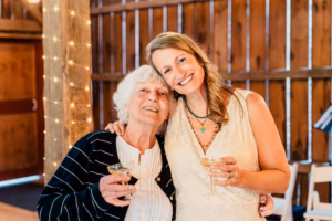 Two women stand closely together in a wooden room, smiling and holding glasses of champagne, with string lights wrapped around a nearby post.