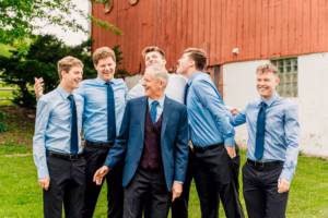 Six men, dressed in blue shirts and ties, stand and laugh together outside in front of a red wooden building on a grassy lawn.