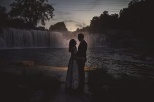A couple stands facing each other near a waterfall at dusk, silhouetted against the water and twilight sky.