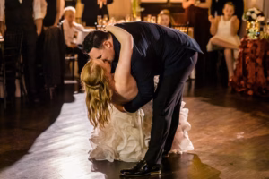 A groom in a dark suit dips and kisses his bride in a white dress on a dance floor during their wedding reception, with guests seated in the background.