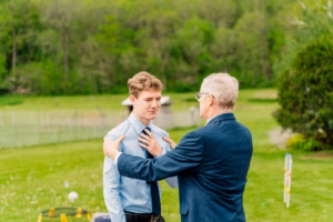 An older man in a suit holds a younger man's shoulders outdoors on a grassy field, with trees and sports equipment visible in the background.