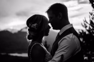 A bride and groom stand close together in silhouette, facing each other outdoors at sunset, with mountains and trees in the background.