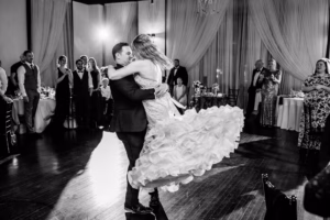 A newlywed couple shares their first dance as guests watch, with the bride’s dress swirling and the groom lifting her in a formal, dimly lit reception room.