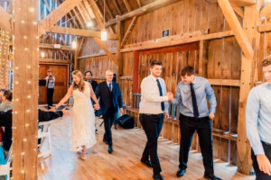 People in formal attire walk and greet each other inside a wooden barn decorated with string lights during an indoor event.