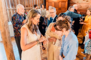 A woman in a white dress shows her ring to two women while a group of people converse in the background in a rustic indoor setting.