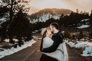 A couple wrapped in a blanket kisses on a paved path with snow, pine trees, and mountains in the background at sunset.