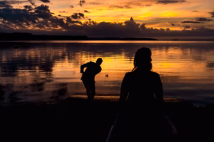 Silhouettes of two people by a lake at sunset; one skips a stone across the water while the other looks on.