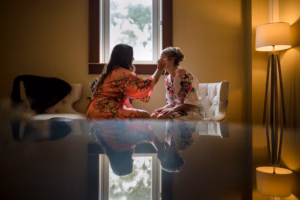 Two women in floral robes sit in a softly lit room; one applies makeup to the other, who is seated and facing her, with their reflection visible on a shiny surface below.
