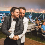 Two men in tuxedos dance together at an outdoor wedding reception under a tent, with seated guests watching in the background.
