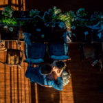 Overhead view of a couple embracing on a wooden floor next to a decorated table with chairs, surrounded by scattered plates, cups, and floral arrangements.