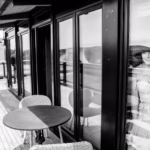A woman in a wedding dress looks out from behind a glass door while a man stands on a wooden terrace outside, facing away.