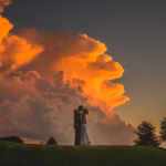 A couple in wedding attire embrace on a grassy hill at sunset, with large orange clouds and trees in the background.