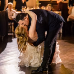 A couple shares a dramatic dip and kiss on the dance floor during a formal event, with guests seated and watching in the background.