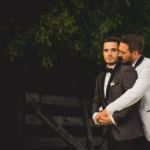 Two men in tuxedos embrace outdoors; one man in a white jacket hugs the other from behind. Green foliage and a wooden fence are in the background.