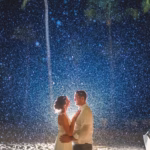 A couple stands together on a sandy beach at night, illuminated from behind, with rain or mist falling around them and palm trees in the background.
