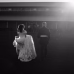 A bride in a wedding dress and a man in formal attire walk toward a large barn-like building at sunset.