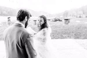 A bride and groom stand outside, facing each other. The bride smiles while holding a piece of paper. The background shows grass, trees, and distant hills.