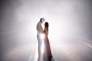 A couple stands facing each other on a foggy road, illuminated from behind by bright white lights, creating a glowing silhouette effect.