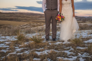 A bride and groom stand side by side outdoors on patchy snow and grass, with the bride holding a colorful bouquet. Their faces are not visible.